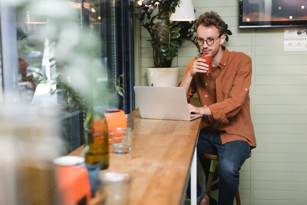 freelancer in glasses looking at laptop and holding cup  - Fotoğraf, Görsel