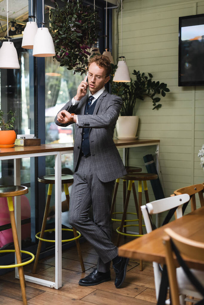 teljes hossza fiatal üzletember formális viselet nézi karóra, miközben beszél okostelefonon kávézóban - Fotó, kép