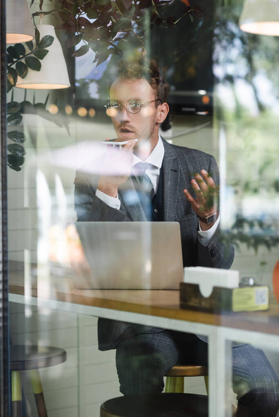 hombre de negocios rizado en gafas de grabación de mensaje de voz cerca de la computadora portátil detrás de la ventana borrosa de la cafetería - Foto, imagen