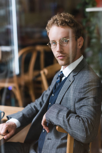 уверенный в себе бизнесмен в очках смотрит в камеру через размытое окно в кафе - Фото, изображение