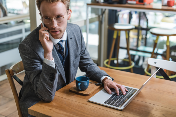 curly businessman in glasses talking on mobile phone near laptop and cup with coffee in cafe - Photo, image
