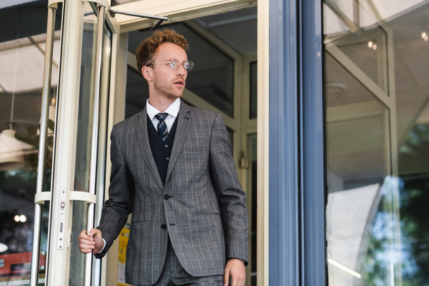 hombre de negocios rizado en gafas y elegante traje abriendo la puerta mientras sale de la cafetería  - Foto, imagen