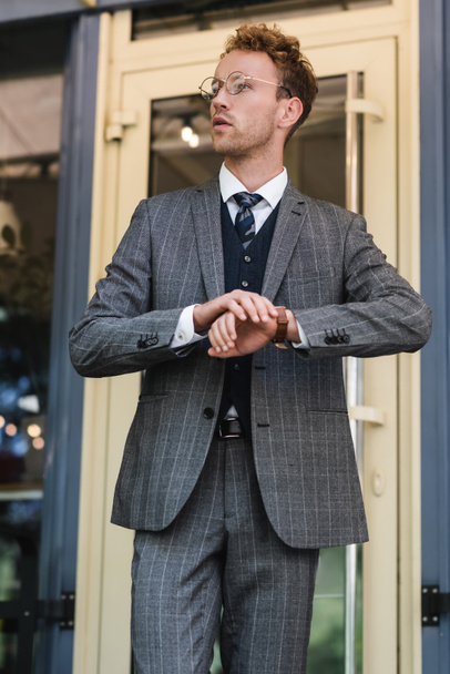 curly businessman in classy suit adjusting wristwatch near cafe entrance - Фото, изображение