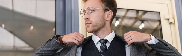 lockiger Geschäftsmann in Brille und edlem Anzug mit Blazer in der Nähe des Caféeingangs, Banner  - Foto, Bild