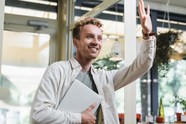 χαρούμενη ελεύθερος επαγγελματίας κρατώντας φορητό υπολογιστή και χαιρετώντας το χέρι κατά την είσοδο cafe  - Φωτογραφία, εικόνα