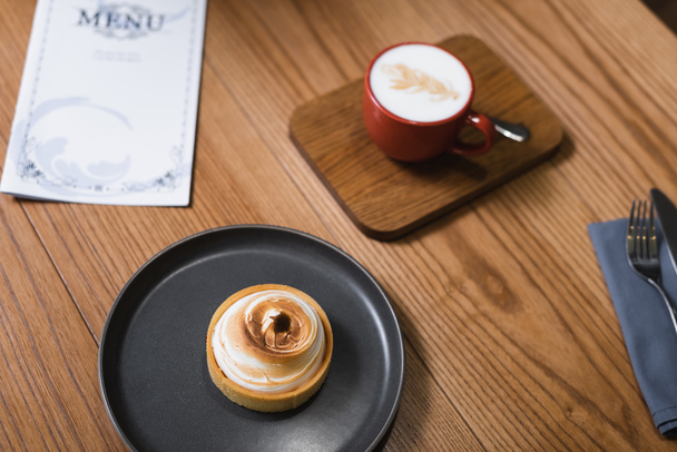 Blick von oben auf schmackhafte Zitronentorte und Cappuccino in der Nähe des Menüs auf dem Holztisch  - Foto, Bild