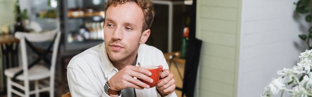 curly man holding cup of cappuccino and looking away in cafe, banner - Photo, image