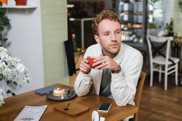 curly man holding cup of cappuccino near gadget and lemon tart while looking away in cafe - Фото, изображение
