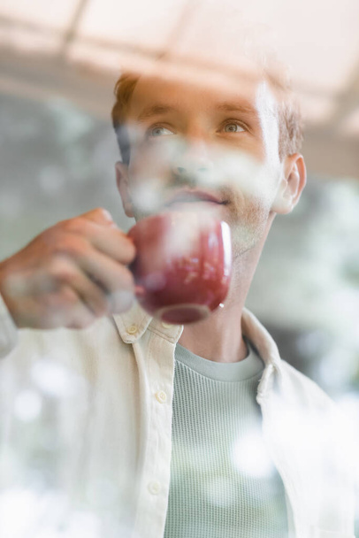νεαρός άνδρας πίνοντας καφέ πίσω από θολό παράθυρο στο καφέ  - Φωτογραφία, εικόνα