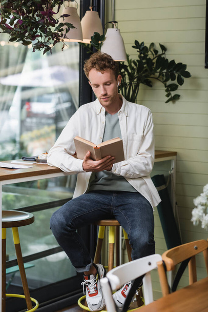 curly man in wireless earphones reading book near menu on table in cafe - Photo, image