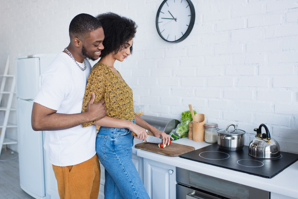 Uśmiechnięty Afrykanin amerykański mężczyzna przytulający dziewczynę krojący sałatkę w kuchni  - Zdjęcie, obraz