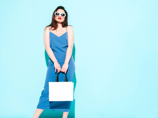 High fashion portrait of young beautiful brunette woman wearing nice blue summer dress.Sexy trendy model posing near blue wall in studio.Fashionable female in sunglasses. Holding shopping bag - Foto, afbeelding