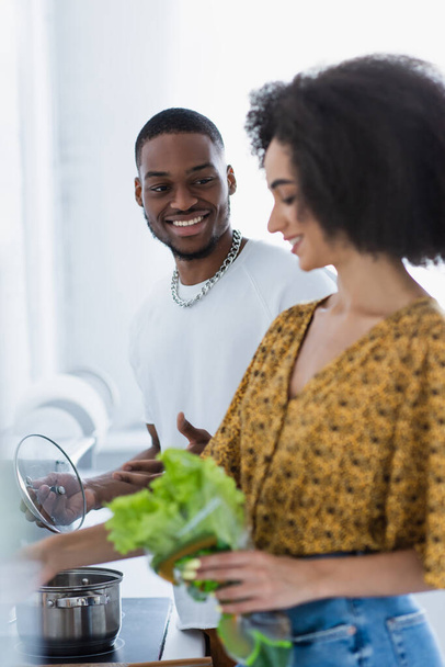 Smiling african american man looking at girlfriend with lettuce on blurred foreground  - Foto, Bild