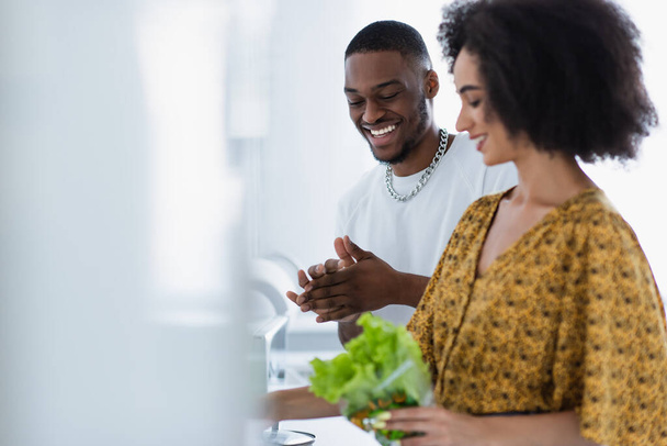 Positive african american man looking at blurred girlfriend with lettuce at home  - Φωτογραφία, εικόνα