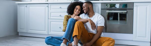 Молода афроамериканська пара посміхається, сидячи на підлозі на кухні, на банері.  - Фото, зображення