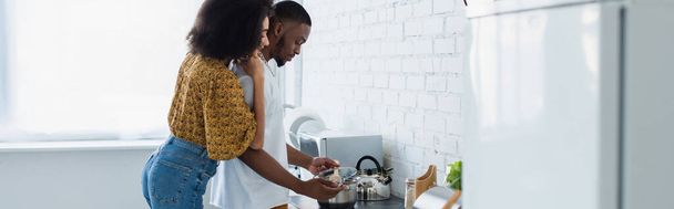 Side view of african american woman hugging boyfriend near stove in kitchen, banner  - Foto, immagini