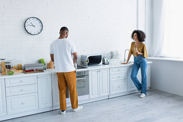 Mujer afroamericana sosteniendo vino mientras su novio cocina en la cocina  - Foto, Imagen