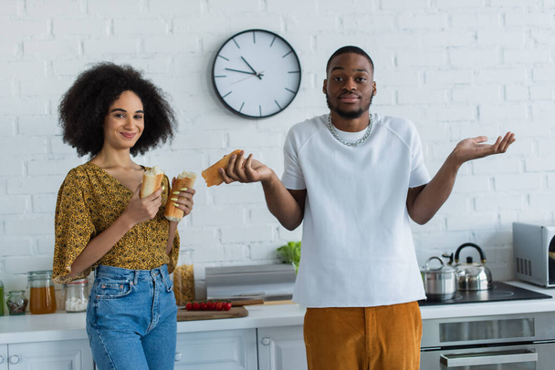 Confused african american man standing near smiling girlfriend with baguette  - Фото, зображення