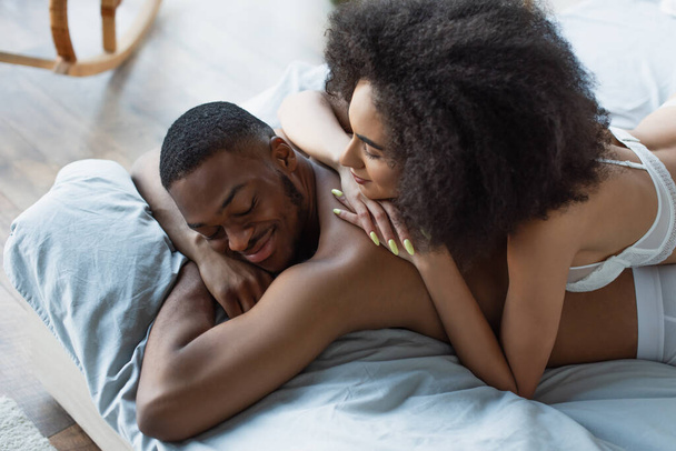 High angle view of african american woman in bra lying on smiling boyfriend in bedroom  - Photo, Image