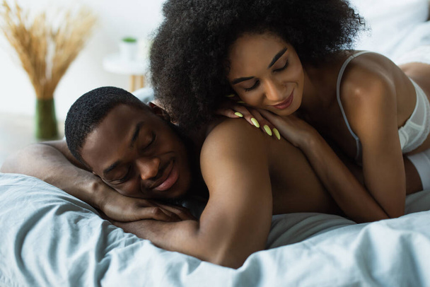 Afroamerikanerin im BH liegt auf Freund mit geschlossenen Augen auf Bett  - Foto, Bild