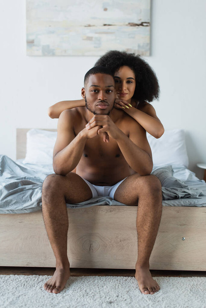 Αφροαμερικανή γυναίκα που κοιτάζει την κάμερα ενώ αγκαλιάζει τον γυμνό φίλο της στο κρεβάτι  - Φωτογραφία, εικόνα