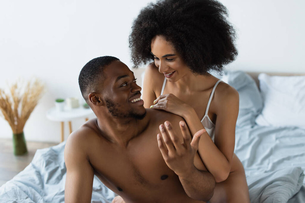 Sütyen giymiş gülümseyen Afro-Amerikan kadın yatakta erkek arkadaşına sarılıyor.  - Fotoğraf, Görsel