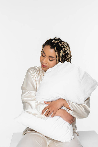 somnolienta y sonriente mujer afroamericana abrazando almohada sobre blanco - Foto, Imagen