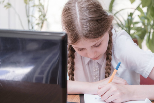 Smart schoolgirl does homework sitting at her desk writes with a pen in her writing notes and uses laptop. She learning language online, doing school tasks at home, listening to lecture - Zdjęcie, obraz