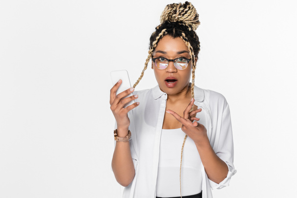 surprised african american businesswoman pointing at smartphone while looking at camera isolated on white - Фото, изображение