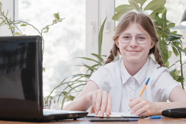 Smart schoolgirl does homework sitting at her desk writes with a pen in her writing notes and uses laptop. She learning language online, doing school tasks at home, listening to lecture - Φωτογραφία, εικόνα
