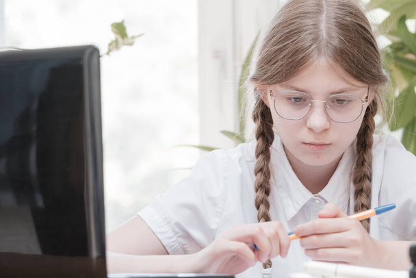 Smart schoolgirl does homework sitting at her desk writes with a pen in her writing notes and uses laptop. She learning language online, doing school tasks at home, listening to lecture - Foto, Bild