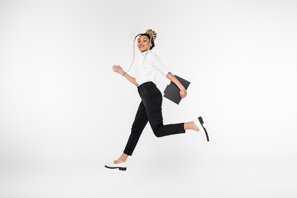 radostná africká americká podnikatelka se usmívá na kameru, zatímco běží v levitaci izolované na bílém - Fotografie, Obrázek