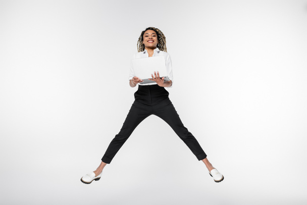 young african american businesswoman smiling at camera while levitating with laptop isolated on white - Photo, Image