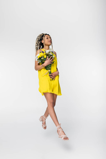 positive african american woman in yellow dress holding flowers while walking in air on white - 写真・画像