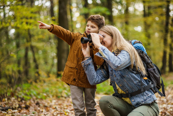 Happy mother and son are hiking in forest. They are watching nature with binoculars. - Foto, afbeelding