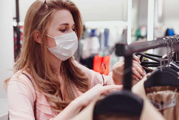 Mooie vrouwelijke klant draagt beschermend gezichtsmasker winkelen in kledingwinkel. Diverse mensen in de modieuze detailhandel, kiezen voor stijlvolle kleding, kleurrijke duurzame merkontwerpen - Foto, afbeelding