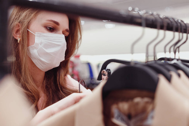 Όμορφη γυναίκα πελάτης φορώντας προστατευτική μάσκα προσώπου ψώνια στο κατάστημα ρούχων. Διαφορετικοί άνθρωποι στο κατάστημα μόδας, επιλέγοντας κομψά ρούχα, πολύχρωμα βιώσιμα σχέδια εμπορικών σημάτων - Φωτογραφία, εικόνα