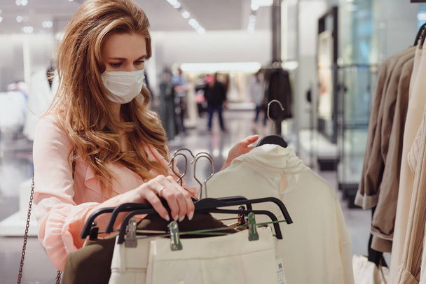Όμορφη γυναίκα πελάτης φορώντας προστατευτική μάσκα προσώπου ψώνια στο κατάστημα ρούχων. Διαφορετικοί άνθρωποι στο κατάστημα μόδας, επιλέγοντας κομψά ρούχα, πολύχρωμα βιώσιμα σχέδια εμπορικών σημάτων - Φωτογραφία, εικόνα