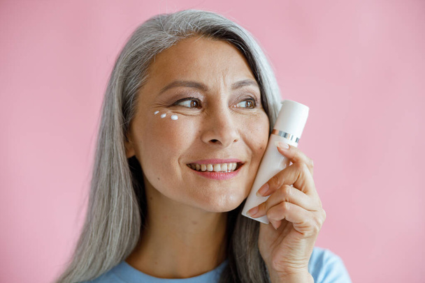 Positive middle aged Asian model with cream drops under eyes holds bottle on pink background - Photo, image