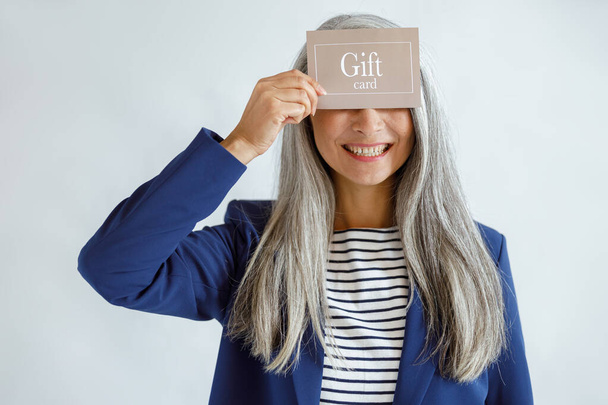 Усміхнена зріла дама з вільним волоссям тримає подарункову картку, позує на світло-сірому фоні
 - Фото, зображення