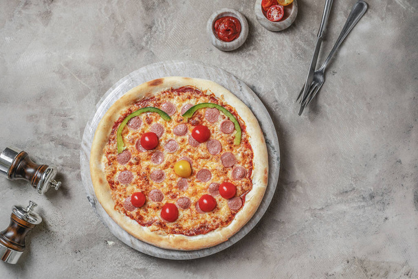Sabrosa pizza con salchicha, mozzarella para niños con sonrisa en bandeja redonda de mármol. Decorado sobre fondo de piedra gris con ingredientes de pizza  - Foto, imagen