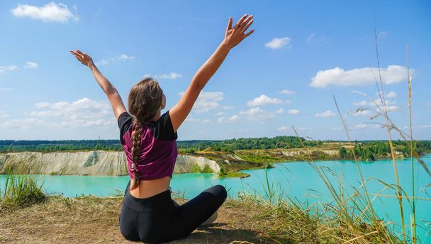 Young woman in leggings and a top with her hands raised up sits in a lotus position on the edge of a chalk quarry on a sunny summer day, Belarus. Concept of freedom, sport - Photo, Image