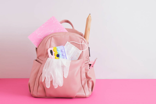 leather pink backpack on gray background, school ladies backpack with big head, smartphone with pop it case, large pencil and notebook, back to school concept, face mask and gloves, lockdown at school - Foto, Imagen