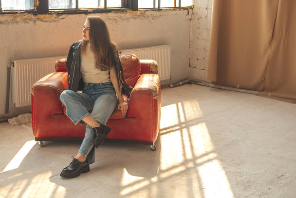 Jeune fille assise sur un vieux fauteuil en cuir rouge à l'intérieur du studio, vêtements décontractés et urbains et de la mode  - Photo, image