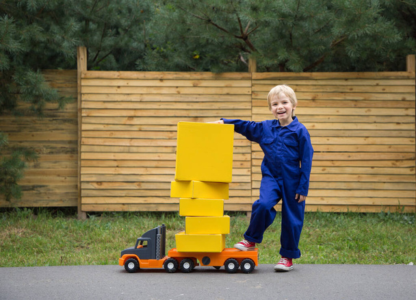 5-летний мальчик в синем комбинезоне стоит рядом с большой игрушечной машиной - грузовиком, загруженным желтыми картонными коробками. Маленький курьер на работе. Доставка посылки, маленький почтальон. быть как папа. игры для мальчиков - Фото, изображение