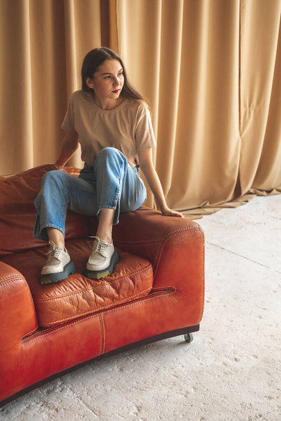 Casual retro e menina adolescente vintage em estúdio. Menina sentada em poltrona vermelha retro em jeans jeans jeans e tênis urbanos, conceito de moda  - Foto, Imagem