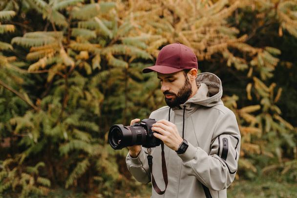 stylový vousatý fotograf s fotoaparátem v ruce pořídí snímek v podzimním parku - Fotografie, Obrázek