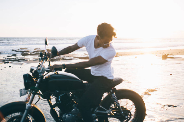 Ethnic curly haired young male motorcyclist in stylish outfit and eyeglasses riding motorbike while looking dowm at sunny lonely seaside - Фото, зображення