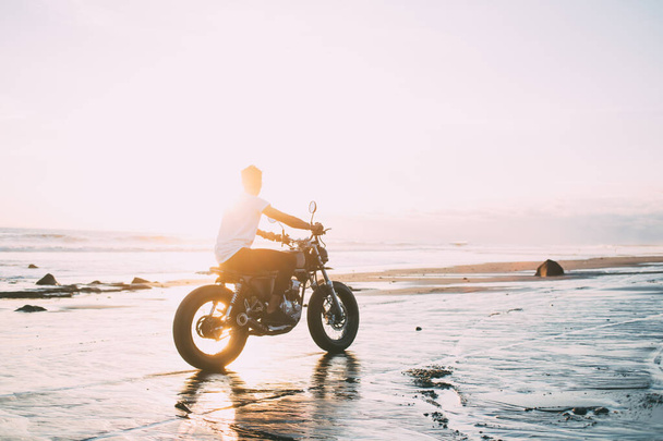 Side view of carefree male motorcyclist in casual clothes watching amazing sunset while riding bike along seashore in back lit - Foto, imagen