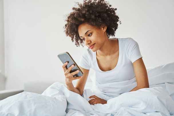 Žena s chytrým telefonem v ruce sedí v posteli - Fotografie, Obrázek